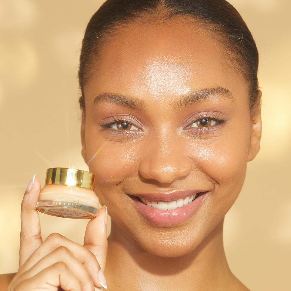 A close-up of a person smiling holding the Miracle Eye Cream Bottle